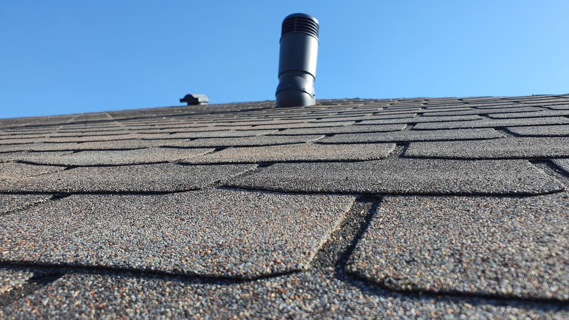 Asphalt shingle roof with a black ventilation pipe against blue sky